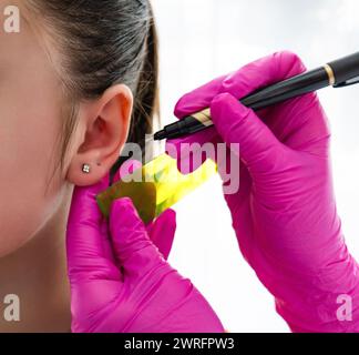 Les mains du cosmétologue dans des gants stériles roses tiennent une règle et un stylo pour mesurer la place de l'oreille d'une patiente pour percer la boucle d'oreille. Haute qualité p Banque D'Images