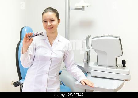 Une femme en manteau blanc avec des manches bleues électriques sourit tout en tenant une paire de lunettes. Son geste professionnel vient de son travail en ophtalmolo Banque D'Images