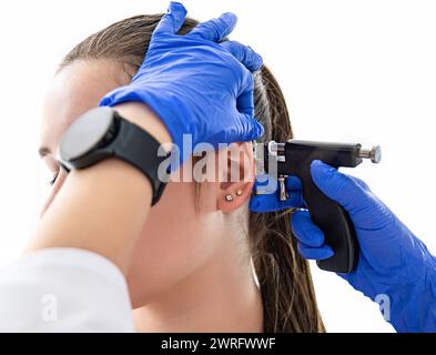 Un médecin portant des gants médicaux bleus stériles perce les oreilles d'une jeune fille dans le cabinet médical avec un pistolet de perçage. Photo de haute qualité Banque D'Images