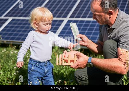 Fils et père économisent de l'argent ensemble, mettant de l'argent dans la tirelire sur fond de panneaux solaires. Papa apprend à petit enfant comment économiser de l'argent pour l'avenir. Concept d'investissement dans les énergies renouvelables. Banque D'Images