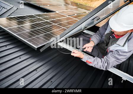 Électricien connectant les câbles tout en installant des panneaux solaires photovoltaïques sur le toit de la maison. Travailleur en casque blanc fixant les câbles de panneau solaire sur un toit. Concept d'énergie alternative et renouvelable. Banque D'Images