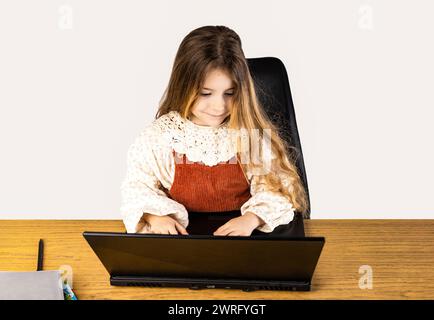 La petite fille est assise à un bureau avec un ordinateur portable sur sa cuisse. Le plancher en bois contraste avec le gadget élégant dans une pochette de protection Banque D'Images