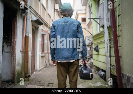 Vue arrière d'un jeune homme à la mode avec lecteur de cassette vintage à la main, errant dans les ruelles de la ville historique. Style de mode rétro moderne tendance des années 90 Banque D'Images