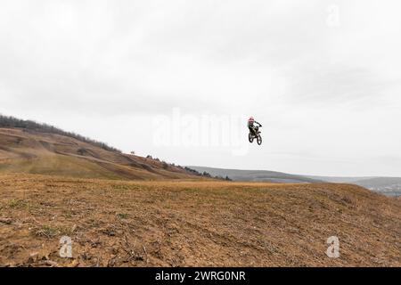 Une personne vole dans le ciel sur un vélo tout terrain au-dessus d'un champ herbeux, avec des nuages et un hélicoptère au loin. Ils mettent en valeur leurs compétences Banque D'Images