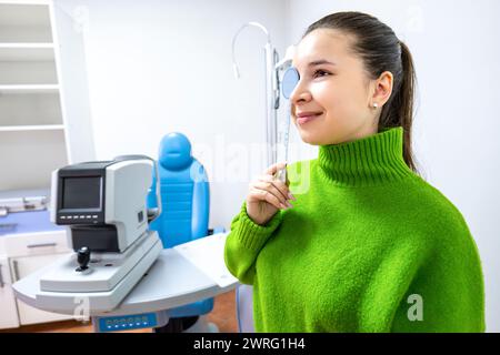 Une femme heureuse dans un pull vert se tient debout devant une machine d'examen de la vue, souriant tandis qu'elle fait des gestes vers l'équipement de bureau sur le bureau Banque D'Images