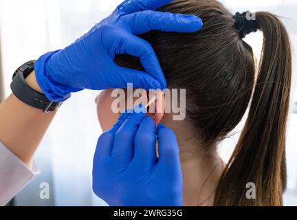 Les mains d'une cosmétologue féminine désinfectant la zone de l'oreille avec un coton-tige, où elle percera les boucles d'oreilles médicinales avec le pistolet. Qual. Élevé Banque D'Images