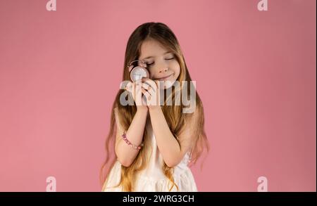 Une petite fille très tendre qui montre des émotions positives envers l'horloge rose, la petite fille vêtue d'une jupe tient une horloge dans sa main et s'assoit wi Banque D'Images