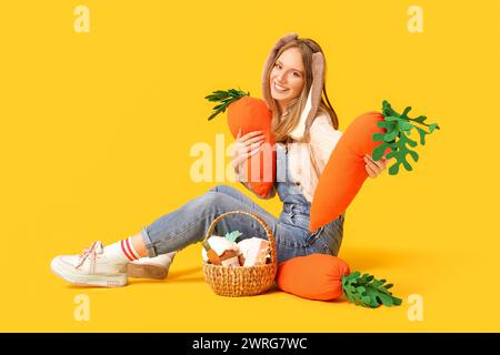 Belle femme aux oreilles de lapin avec des carottes en jouet et panier de Pâques assis sur fond jaune Banque D'Images