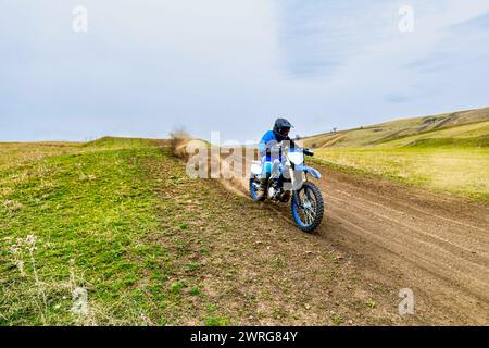 Une personne accélère sur une moto avec un casque de course sur un chemin de terre, dégageant la poussière et l'herbe pendant que le pneu en caoutchouc adhère au sol Banque D'Images