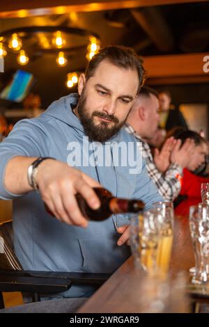 Un jeune homme beau avec une barbe versant de la bière de la bouteille dans des verres pour des amis. Le concept d'une fête de pub. Banque D'Images