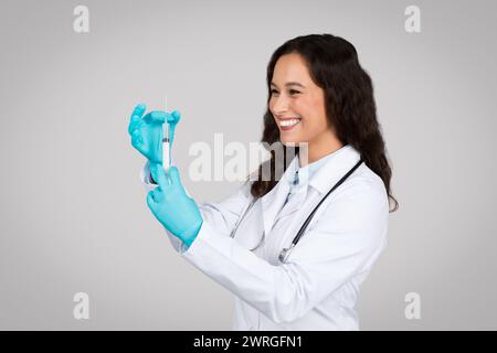 Jeune femme joyeuse médecin en blouse de laboratoire blanche et gants préparant soigneusement la seringue pour la vaccination Banque D'Images