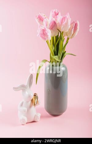 Tulipes roses dans un vase et figurine blanche de lapin de Pâques sur fond rose pastel Banque D'Images