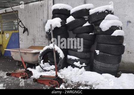 Une pile de vieux pneus usagés et plusieurs crics alignés devant un atelier. La neige leur tombait dessus. Banque D'Images