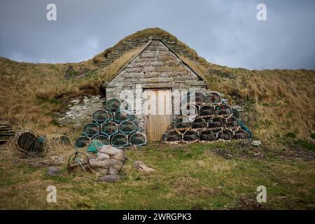 Chalet en pierre / hangar à l'entrée du port de Sinclairs Bay avec des casiers de homard et de crabe empilés à l'extérieur par une journée ensoleillée en Écosse rurale, prêt pour la pêche Banque D'Images