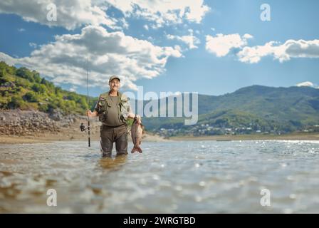 Pêcheur en uniforme debout à l'intérieur d'un lac de montagne et tenant un poisson Banque D'Images