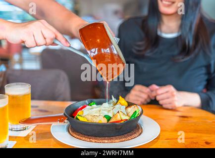 Concentrez-vous sur les mains d'un serveur de restaurant tenant une poêle en céramique et versant du fromage fondu sur un plat en fonte rempli de croûtons de pain et Banque D'Images