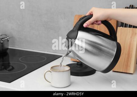 Femme versant de l'eau chaude de la bouilloire électrique dans la tasse dans la cuisine, gros plan Banque D'Images