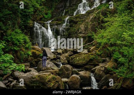 Vue arrière d'une femme randonneuse touristique debout sur des rochers à la chute d'eau de Torc dans le parc national de Killarney, comté de Kerry, Irlande Banque D'Images