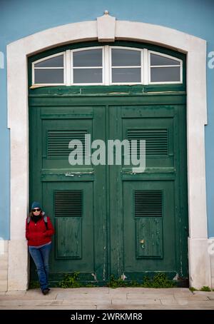 Veste rouge femme s'appuie contre vibrant vert profond vintage porte en bois blanc cadre en pierre bleu mur extérieur Banque D'Images