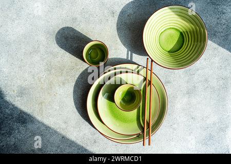 Vue de dessus d'une table élégante avec des plats en céramique verte, des bols et une tasse avec des baguettes brunes, le tout baigné par la lumière naturelle du soleil avec ombre Banque D'Images