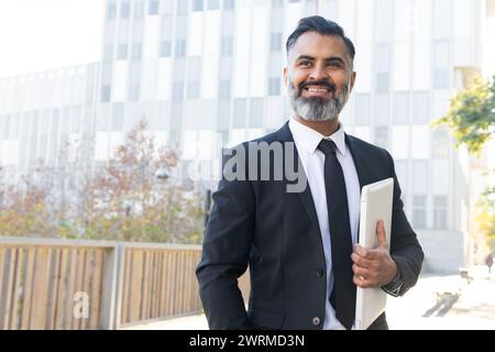 Homme professionnel en costume tenant un ordinateur portable sourit en toute confiance à l'extérieur d'un immeuble de bureaux. Banque D'Images