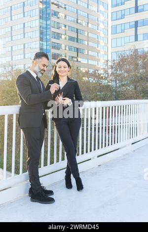 Un homme et une femme en tenue d'affaires sont engagés dans une conversation joyeuse sur une tablette numérique à l'extérieur des immeubles de bureaux. Banque D'Images