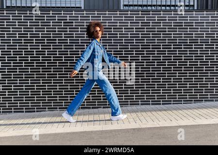 Une femme afro-américaine confiante en plein denim avance, sa pose dynamique contre les lignes graphiques d'un mur de briques Banque D'Images