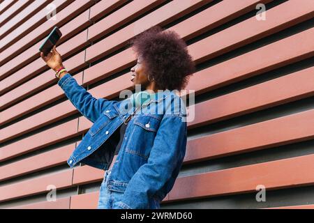 Jeune femme exubérante capturant un moment selfie, son style mis en valeur par une veste en denim sur un fond rayé Banque D'Images