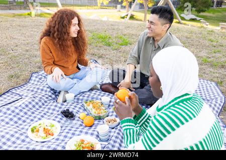 Un groupe diversifié d'amis se réunit pour un pique-nique sur une couverture à carreaux dans le parc, partageant sourires, nourriture et conversation. Banque D'Images