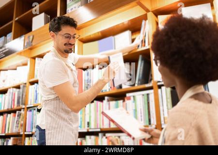 Un jeune homme tend un livre à une femme afro-américaine au milieu des allées d'une bibliothèque bien garnie Banque D'Images