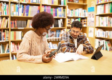 Deux jeunes étudiants participent à une session d’étude dans une bibliothèque, avec des livres, une tablette numérique et un smartphone Banque D'Images