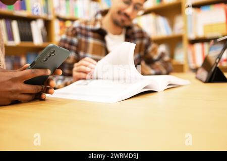 Gros plan de la main d'une personne tenant un smartphone, avec une autre personne étudiant en arrière-plan dans une bibliothèque. Banque D'Images