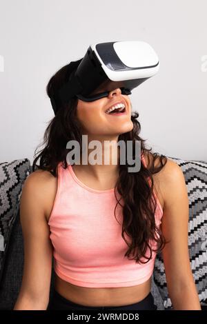 Une jeune femme adulte hispanique joyeuse, porte un casque de réalité virtuelle assis dans un canapé à la maison Banque D'Images