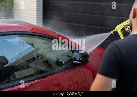 Une personne utilise un jet d'eau à haute pression pour nettoyer la vitre latérale d'une voiture rouge brillante Banque D'Images