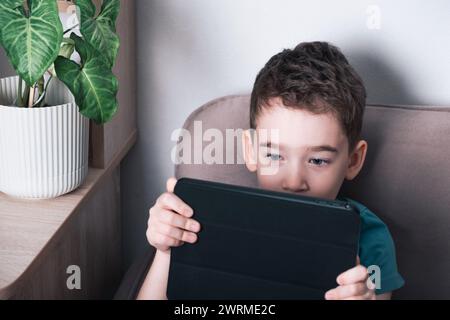 Un enfant concentré s'assoit confortablement, profondément engagé avec une tablette numérique, soulignant l'influence de la technologie moderne sur les jeunes. Banque D'Images