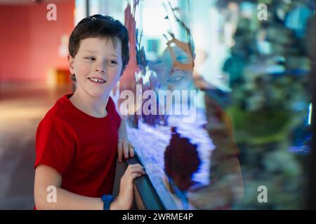 Un garçon portant une chemise rouge et une montre sourit à une exposition interactive sur l'eau reflétant son image dans un musée des sciences. Banque D'Images