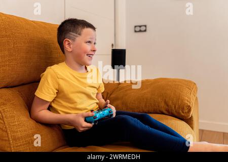 Un jeune garçon s'assoit confortablement sur un canapé aux tons chauds, jouant joyeusement avec une manette de jeu vidéo, affichant un sourire de plaisir authentique et engagé Banque D'Images