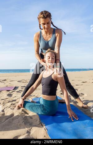 Un instructeur assiste un élève dans une pose de yoga sur la plage avec un ciel dégagé au coucher du soleil. Banque D'Images