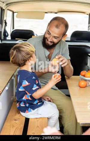Un père et un enfant souriants partagent un moment ludique en épluchant des oranges dans un confortable van vintage, reflétant une expérience insouciante et solidaire. Banque D'Images