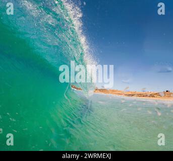 Un gros plan époustouflant d'une vague turquoise translucide se courbant magnifiquement sur un ciel bleu clair, avec le soleil illuminant les textures de l'eau Banque D'Images
