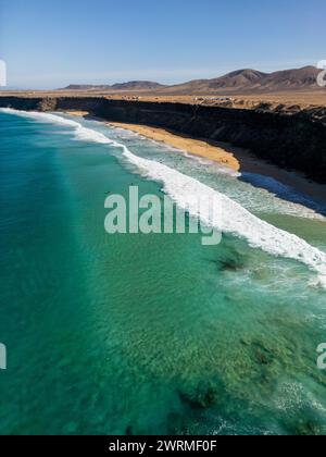 Tir par drone à grand angle capturant les eaux turquoises claires, les vagues ondulantes et la plage de sable le long des falaises accidentées de Fuerteventura. Banque D'Images