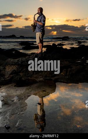 Vue de côté d'un homme se tient sur une plage rocheuse jouant du saxophone au crépuscule, avec un ciel coloré de coucher de soleil et son reflet dans un bassin de marée. Banque D'Images