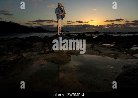Un homme se tient pieds nus sur les rochers côtiers, jouant du saxophone sur un magnifique coucher de soleil en toile de fond, évoquant une atmosphère calme et créative. Banque D'Images