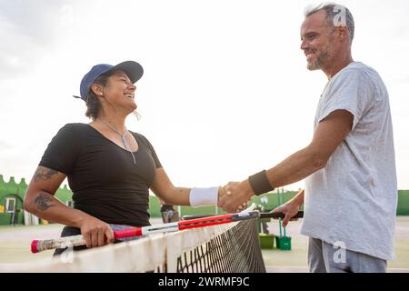 Deux joueurs de tennis joyeux, un homme et une femme, serrent la main sur le filet après le match, mettant en valeur l'esprit sportif et la joie. Banque D'Images