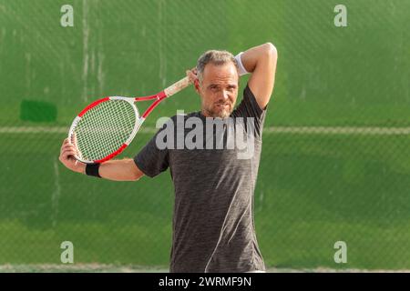 Un homme mature fait une pause sur le court de tennis, regardant loin de la caméra, tenant sa raquette et essuyant la sueur de son front. Banque D'Images