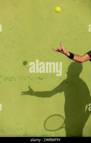 Bras d'homme recadré méconnaissable lançant une balle de tennis en l'air, se préparant à servir, par une journée ensoleillée avec son ombre visible sur le court. Banque D'Images