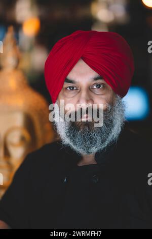 Un homme sérieux avec un turban rouge et une barbe entièrement grise dans un fond ambiant flou Banque D'Images