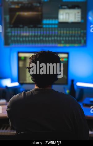 Un producteur de musique se concentre sur le mixage de pistes dans un studio sonore faiblement éclairé entouré d'un équipement audio professionnel et d'un éclairage ambiant bleu. Banque D'Images
