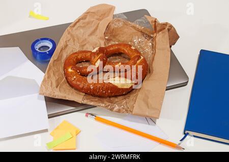 Une collation de bretzel fraîchement cuit sur du papier kraft au milieu des fournitures de bureau, y compris un cahier, un crayon et des notes adhésives. Banque D'Images