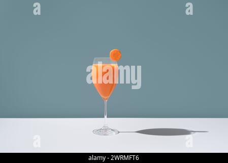 Un élégant verre rempli de jus de carotte orange vif, garni d'une tranche de carotte fraîche placée sur le bord, sur un fond bleu doux. Banque D'Images